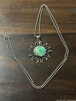 Sterling silver necklace with sun-shaped pendant and central blue-green stone on a ball link chain against brown background.