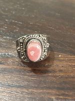 Close-up photo of the sterling silver ring on a wooden surface showing the pink oval stone and detailed silver bead and rope design around the bezel.