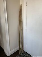Photo showing all ten bamboo sticks leaning against a corner of the room, varying heights visible, natural light tan color and straight bamboo stalks