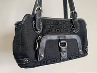 Front and side view of black handbag showing geometric pattern fabric, exterior zip pocket with leather and buckle detail, double handles with silver rings.