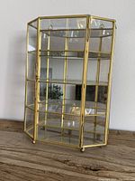 Small brass and glass curio cabinet standing on a wooden surface. Shows front and side glass panels, brass framework, glass shelf and small round feet.