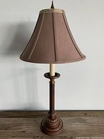 Brown metal lamp with detailed base and brown trumpet-shaped fabric shade, turned off, sitting on wooden surface.