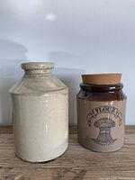 Photo showing two ceramic crocks side by side on a wooden surface against a white wall. The smaller crock is the flour jar with a cork lid and printed wheat sheaf design. The larger beige crockpot is plain with a pale cream color.