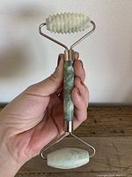 Hand holding jade facial roller showing full length, textured roller on top, smooth roller bottom, metal frame, and polished green jade handle.