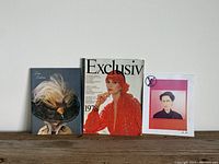 Three fashion magazines displayed upright: Exclusiv 1978 edition, True Fashion magazine with feather hat cover, and Louis Vuitton program with pink border portrait.
