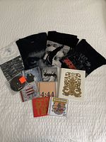 Lot displayed on bed: six concert t-shirts folded, seven CDs in cases arranged below, and one hardcover book with white and gold cover.