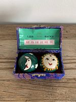 Open fabric box with sun and moon iron meditation balls inside beside printed green label.
