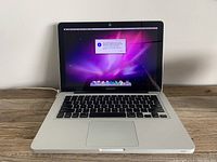 Front view of Apple MacBook Pro laptop powered on showing desktop and alert message on screen. Laptop is open and placed on a wooden surface with power cord connected.