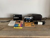 Two vintage Kodak cameras, Kodak Teledisk and Instamatic X-15, with manuals, film, pouch and cameras with straps on wooden surface.