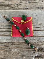 Three-piece jewelry set displayed with red pouch with yellow trim on wooden table. Includes bracelet, ring, and pendant with oval green stones set in metal.