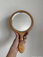 Front view of handheld mirror showing round beveled glass surface and birds eye maple wood frame and handle held in hand