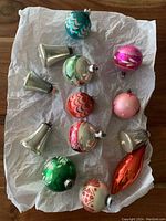 Photo showing 13 vintage glass Christmas ornaments of various shapes and colors laid on white tissue paper including silver bells, spherical ornaments in pink, red, green, blue-green with different painted patterns.