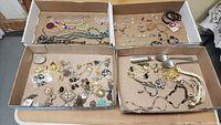 Top view of four cardboard trays filled with various vintage costume jewelry pieces, showing the range of items included such as necklaces, brooches, bracelets, and pins.
