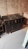 Full view of the antique black steamer trunk showing riveted metal edges and front locks, placed on a concrete floor.