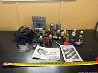 Full view of the airbrush paints, accessories, airbrush unit, and manuals on a black surface with a yellow measuring tape in front for scale.
