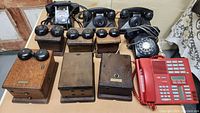 Lot of old telephones and wooden ring boxes arranged on a table, showing various rotary dial phones, keypad phone, and six wooden box units with bells.