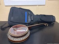 Wide shot showing the banjo with circular drum, wooden neck, metal tuning pegs, and black gig bag with blue zipper accents, placed flat on a black surface.