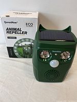 Front photo of green QueenMew animal repeller showing solar panel on top, two LED lights, motion sensor, and speaker grille.