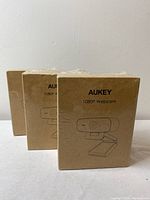 Three sealed brown boxes of Aukey 1080P webcams shown upright with sealed plastic wrapping, illustrating the packaging and quantity in lot.