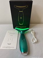 Beauty Neck Instrument device with its black box, USB charging cable, and instruction manual laid out on table.