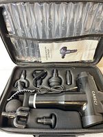 Open carrying case showing massager, interchangeable heads, charging cable, and instruction manual.