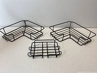 Three black metal shower caddy pieces: two corner baskets and one soap dish placed on a flat surface.