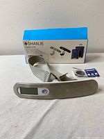 Digital luggage scale with LCD display, strap, original box and CR2032 battery on white surface against white background