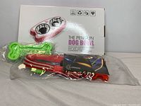 Penguin Dog Bowl box with green bone-shaped chew toy attached and red dog collar in packaging