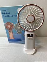 White folding handheld fan with digital display on handle, standing upright with attached base stand in front of its packaging box.