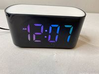 Front view of black LED alarm clock showing digital time display (12:07 AM) with blue and purple numbers.