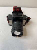 Front view of the LED headlamp showing the large central lens with lens cover, two small circular LED lights above, and two rectangular LED light panels on either side. Adjustable head strap visible on sides.