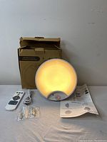 LED lamp glowing warm white light, with remote control, charging cable, mounting screws, anchors, and manuals displayed in front of the opened box