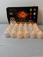 24 LED small tea candles arranged in rows on a table with original packaging box behind them showing colorful LED candle options and quantity information.