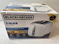 Front and top view of the boxed Black & Decker 2-slice toaster showing product image, brand logo, and key features including extra-wide slots and high-lift lever.