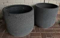 Two large round black resin flower pots with textured surfaces, shown side-by-side on a brick floor against a white brick wall.