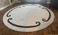 Full view of round rug showing beige field and black scroll border on hardwood floor