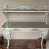Front view of white cast iron side table against a brick wall showing glass top shelf and decorative scroll and floral pattern on the front apron and upper shelf.