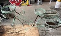 Full side view of turquoise metal decorative bicycle plant holder with scrollwork and three planter baskets attached, showing overall structure and size.