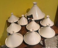 Nine white ceramic tajines (one large, eight small) arranged on wooden table against yellow wall