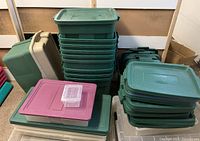 Multiple stacks of Rubbermaid totes and lids in green, beige, clear, and pink colors on garage floor, showing a range of sizes and mostly lids.