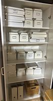 White storage cabinet filled with multiple white storage boxes with metal corner reinforcements, arranged neatly on shelves. Bottom shelf contains a natural-colored rectangular wicker basket and two white magazine holders.