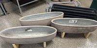 Three concrete stone planters with oval shape and wooden legs; four black rectangular plastic planters with brackets behind them on a concrete floor.
