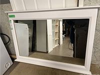 Full view of the large horizontal mirror showing the white frame with trim detail and clear reflective glass, placed on the floor leaning against a wall.