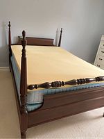 Full-sized wooden bed frame with decorative posts and mattress topped with yellow memory foam layers shown from an angled perspective.