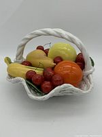 Porcelain fruit basket viewed from the side showing colorful fruit figurines including cherries, banana, orange, and pear in a white twisted wicker-style basket with handle.
