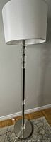 Full view of floor lamp showing white shade and stainless steel pole with glass accents on a wood floor with grey wall.