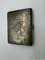Closed metal cigarette case showing embossed scenic design of buildings and trees