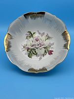 Top view showing round scalloped porcelain bowl with central floral rose painting and gold accent trim on edges against blue background.