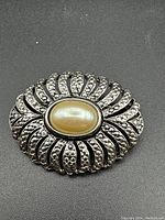 Front view of vintage brooch showing oval faux pearl center and textured silver-toned metal pattern radiating outward.