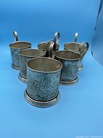 Six silver-tone metal tea glass holders (Podstakannik), embossed with horse and troika scenes, arranged on blue background.